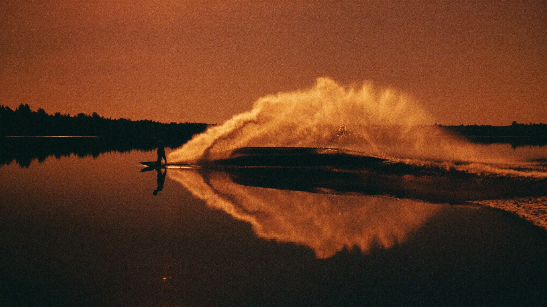 Wakesurfer carving a glass lake at dusk, golden spray